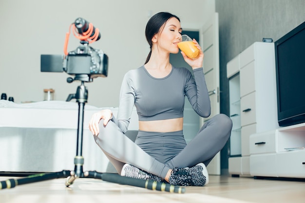 Beautiful young lady in sporty clothes sitting on the floor in lotus pose with a glass of juice Modern camera with a microphone on a tripod