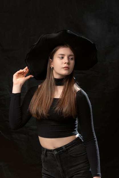 Beautiful young lady wears black clothes and widebrimmed hat Portrait of young model on black background Vertical frame