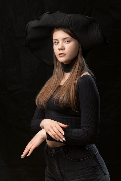 Beautiful young lady wears black clothes and widebrimmed hat Portrait of young modern aristocrat on black background Vertical frame