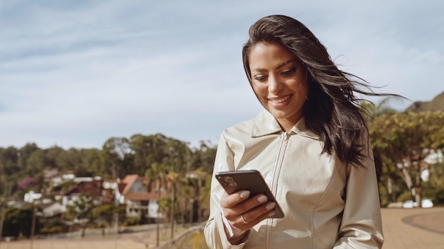 Beautiful young Latin woman using smartphone standing on city street Portrait of Latina smiling female using mobile phone