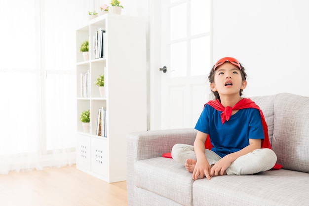 beautiful young little kid children girl play as superhero and sitting on living room sofa couch relaxing daydreaming at home.