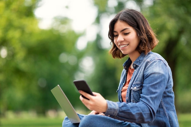 Beautiful Young Middle Eastern Woman Messaging On Smartphone And Using Laptop Outdoors