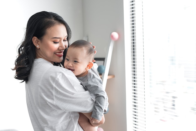 Beautiful young mom is playing with her cute baby and smiling while standing near the window at home