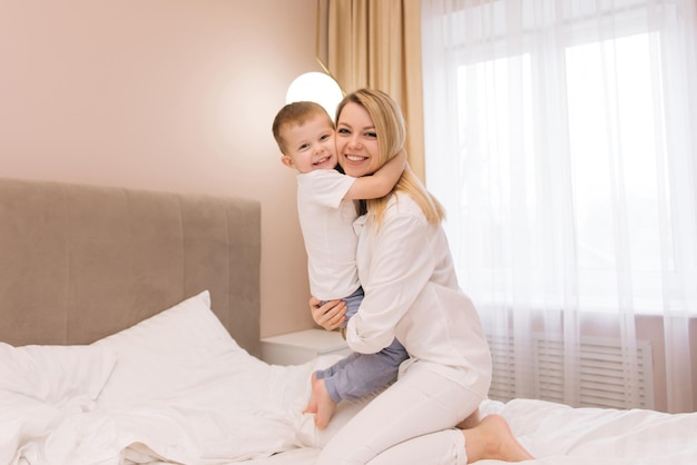 Beautiful young mom playing and laughing with her baby smiling son in the bedroom bed happy family