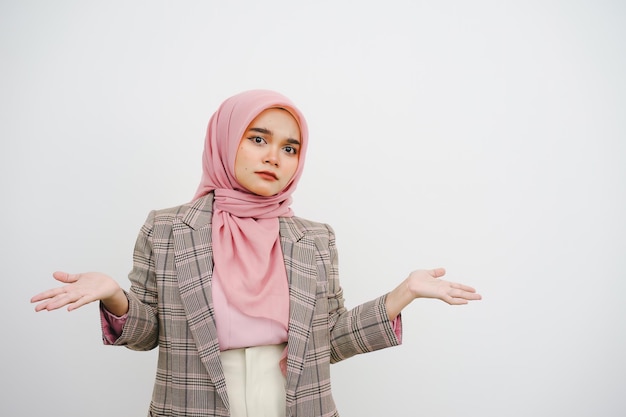 Beautiful young muslim businesswoman pink hijab shrugging shoulders looking puzzled have no idea standing questioned and unaware I don't know why spread hands isolated on white background