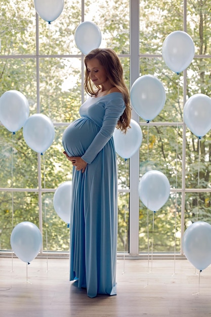 Beautiful young pregnant woman in blue long dress with blue balloons stands at the big window