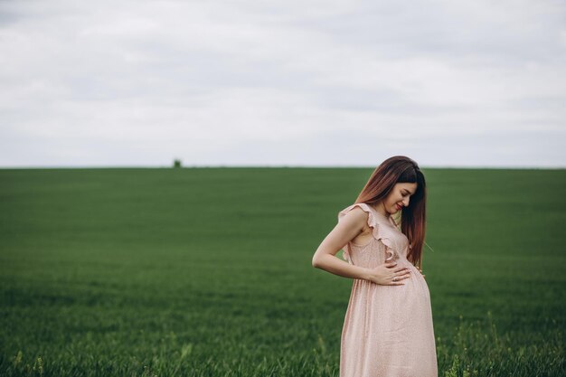 A beautiful young pregnant woman in the hat stands on a green field leaning against a haystack Sunny summer day She is happy