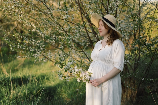 A beautiful young pregnant woman in a white dress walks in the spring garden