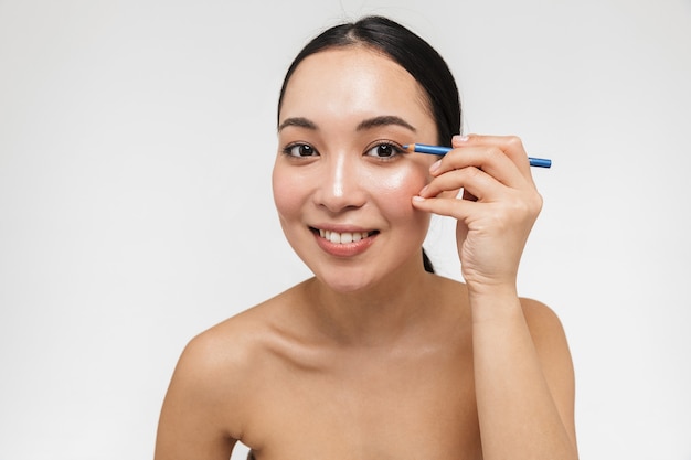 beautiful young pretty asian woman with healthy skin posing naked isolated over white wall holding eye pencil.