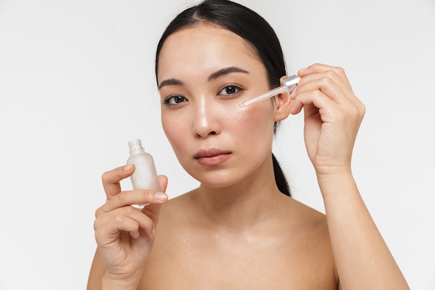 beautiful young pretty asian woman with healthy skin posing naked isolated over white wall take care of her skin with cosmetics.