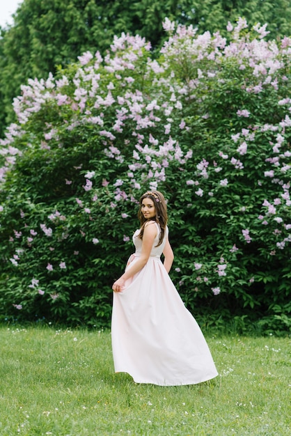 A beautiful young pretty girl with long hair loose stands near a blooming lilac Bush in a long pale pink dress. She looks back and goes forward happy