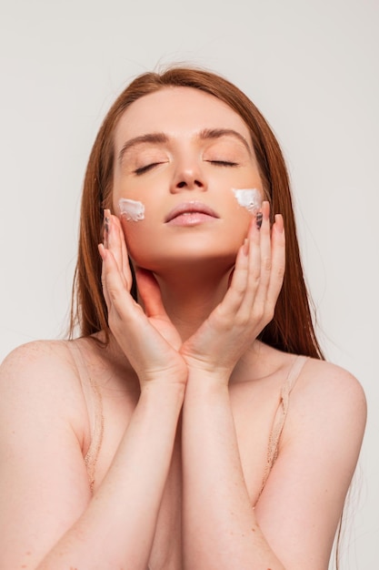 Beautiful young redhead woman in a beige bra does skin care and applies cream on her face