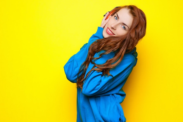 Beautiful young sexy girl DJ in a blue jacket listening music in red headphones on a yellow background and having fun