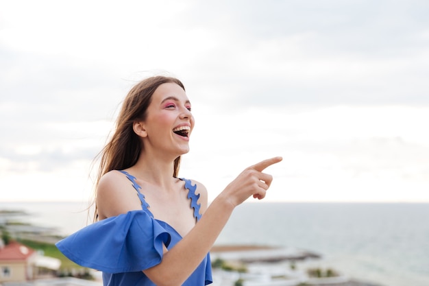 Beautiful young smiling brunette woman standing at the white terrace and pointing finger at something