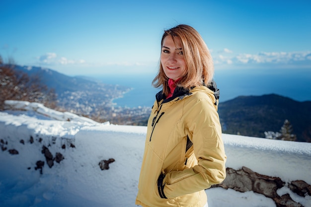 Photo beautiful young smiling girl in her winter warm clothing. on the background of a beautiful winter mountain landscape in nature, on a beautiful observation point in the mountains with sea views