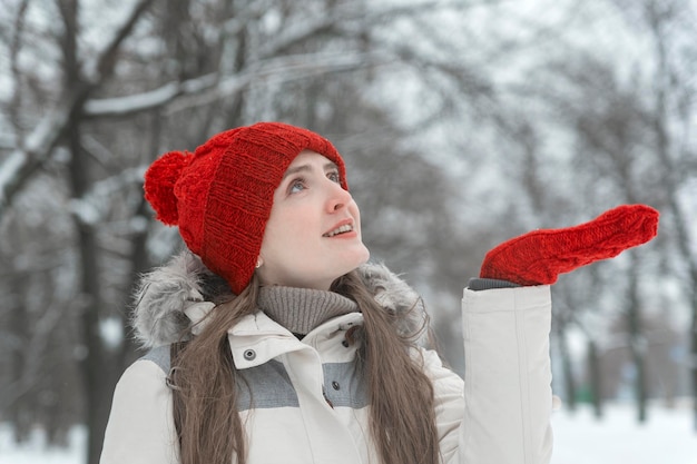 Beautiful young smiling woman in red knitted hat holds out his palm in glove in the woods. Walk through the snow-kept park.