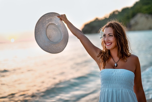 Beautiful young smiling woman in white dress walking on the beach and holding white summer hat.