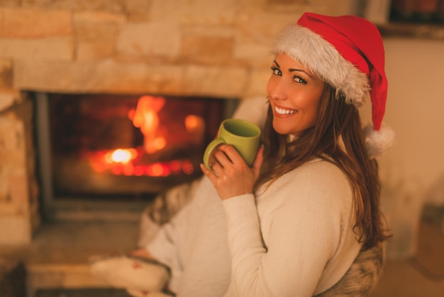 Beautiful young smiling woman with santa hat enjoying a cup of tea by the fireplace. Looking at camera.