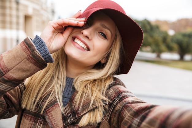 Beautiful young stylish blonde woman wearing a coat walking outdoors, taking a selfie with mobile phone