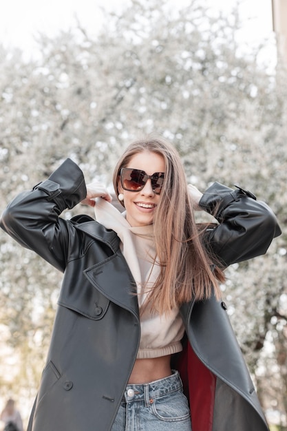 Beautiful young stylish happy girl with a smile in fashionable spring outfit with a long leather coat hoodie and fashion sunglasses is wearing a hood and walking in the park