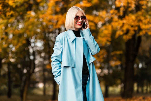 Beautiful young stylish smiling woman with sunglasses in fashionable clothes talking on the phone and walking in the autumn park