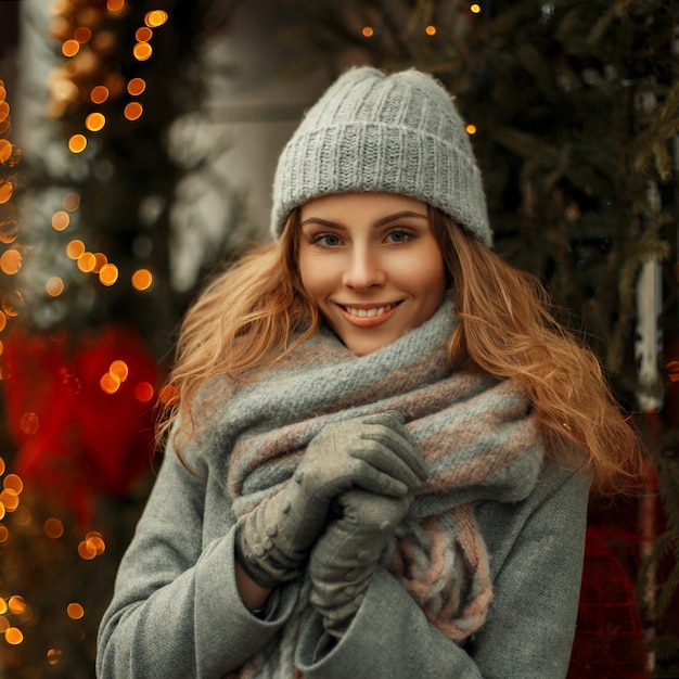 Beautiful young stylish woman smiling in a fashionable knitted hat with a scarf in a trendy gray coat on the street on a background of festive lights on a winter day