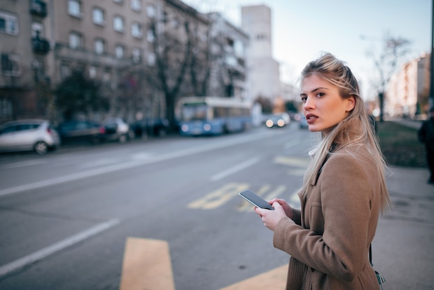 Beautiful young stylish woman using smartpone while waiting for taxi or bus in the city.