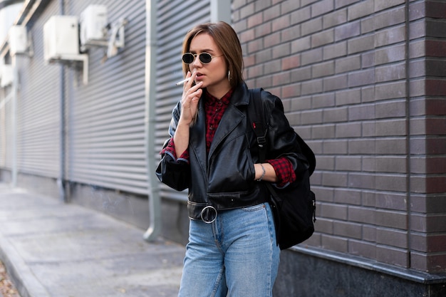 Beautiful young stylishly dressed Caucasian woman smokes cigarette on street Smoking bad habit Nicotine addiction Unhealthy lifestyle Modern woman leather jacket and jeans walking outdoor sunglasses