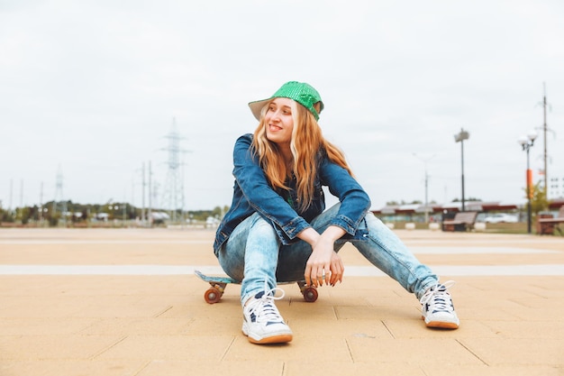 A beautiful young teenage girl in a cap and denim jacket sitting over a skateboard generation z