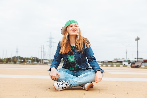 A beautiful young teenage girl in a cap and denim jacket sitting over a skateboard generation z