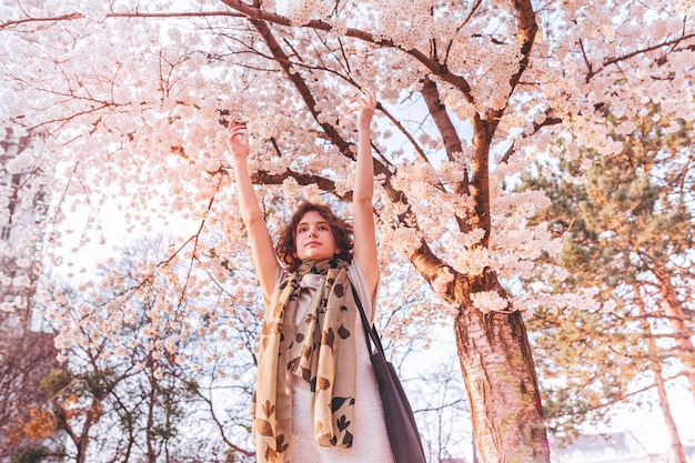 beautiful young teenage girl with brown hair raising hands admires cherry blossoms in sun