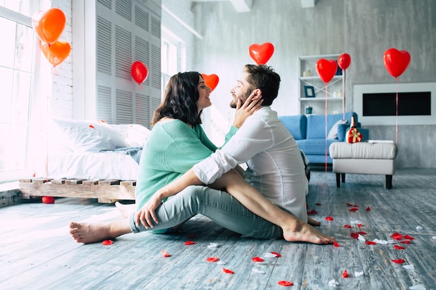 Beautiful young tender lovely couple are kissing and hugging each other while sitting on the floor of bedroom with romantic background. St Valentine's day. Anniversary. Date. Love concept