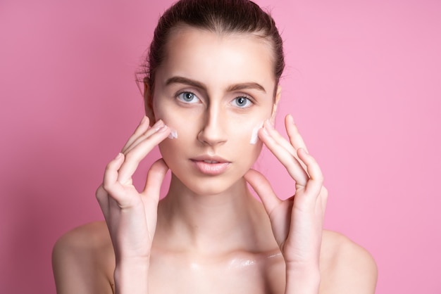 Beautiful young woman applying cream on her face on pink