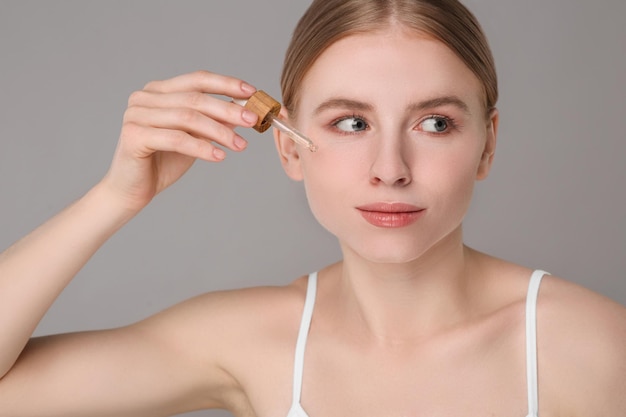 Photo beautiful young woman applying essential oil onto face on grey background