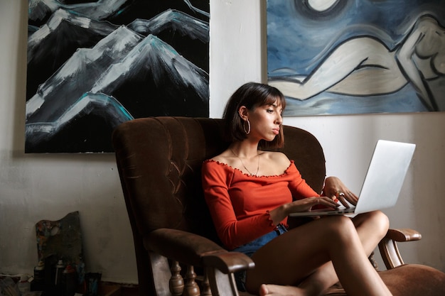 Beautiful young woman artist sitting on chair in the art studio, relaxing, using laptop computer