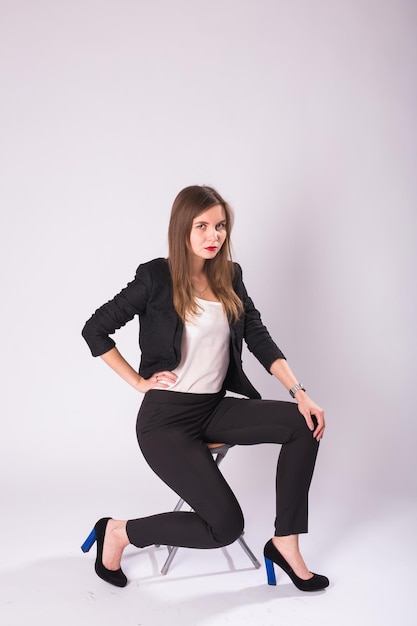 Beautiful young woman in black suit sits on a chair on white background