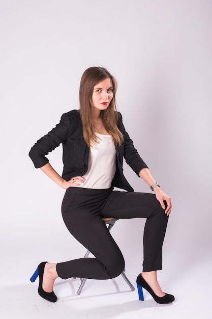 Beautiful young woman in black suit sits on a chair on white background