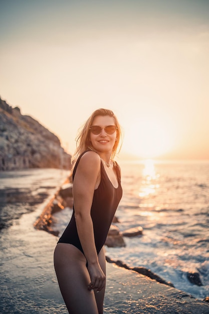 Beautiful young woman in a black swimsuit alone on the beach by the sea at sunset An adult slender girl resting on the ocean shore at sunset Summer vacation concept Selective focus