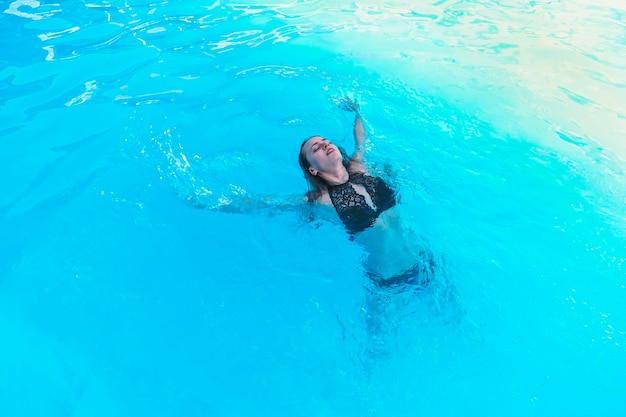 Beautiful young woman in black swimsuit swimming in blue pool on her back Young female swimmer at holiday resort Sport activity health concept smile on face