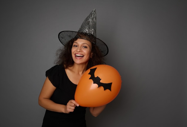 Beautiful young woman in black and wizard hat poses with an orange air balloon with a handmade felt-cut bat, smiles toothy smile looking at camera. Halloween concept on grey background, copy space