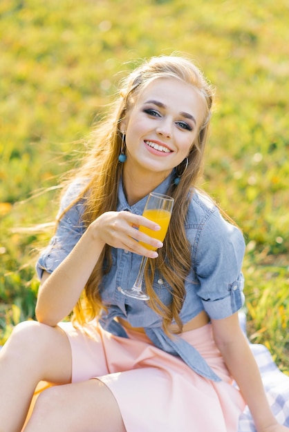 Beautiful young woman in a blue denim shirt and pink skirt in the garden at a picnic holding a glass of juice in her hand