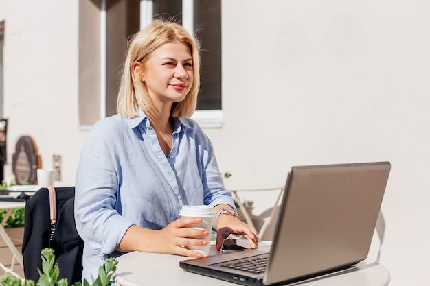 Beautiful young woman in a blue shirt working on a notebook in a cafe and drink coffee Remote work