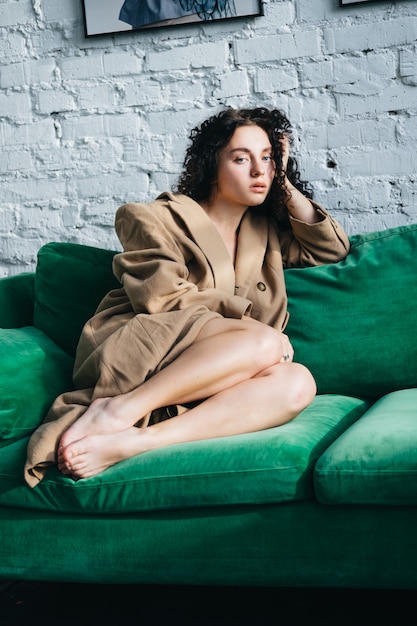 A beautiful young woman in a brown coat is sitting in a room on a green sofa. Interior.