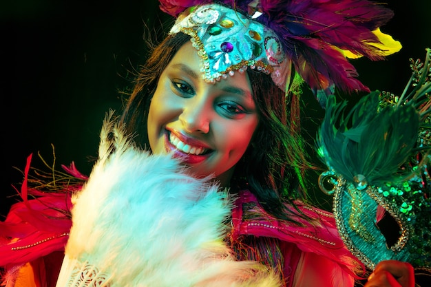 Beautiful young woman in carnival mask and stylish masquerade costume with feathers fan in colorful lights and glow on black background