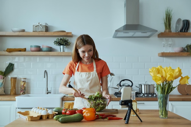 Beautiful young woman cooking and vlogging while standing at the domestic kitchen