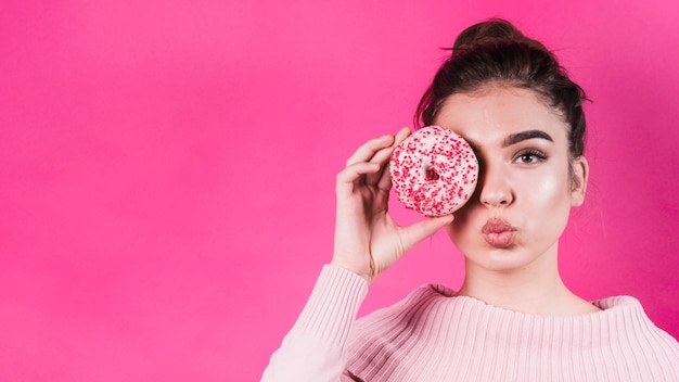 Beautiful young woman covering her eyes with delicious donut covering her eyes against pink background