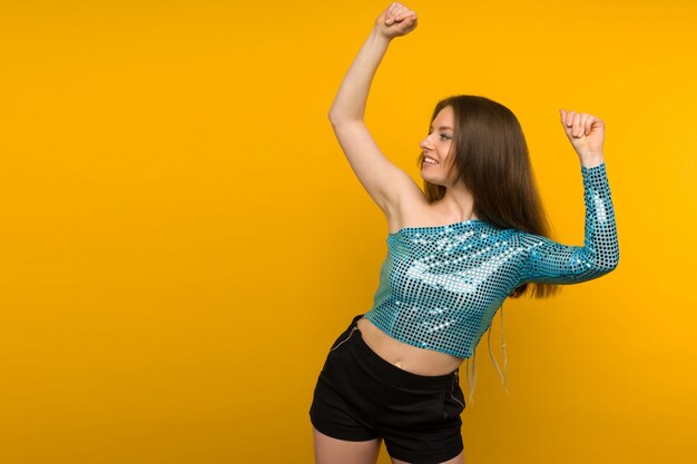 Beautiful young woman dancer in a shiny blue cape posing on a yellow background.