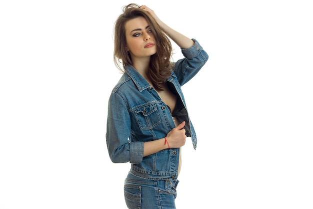 Beautiful young woman in denim clothes holding her hand near the hair and looks toward isolated on white wall