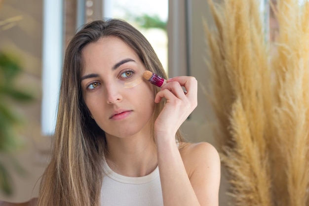 Beautiful young woman doing her makeup in the mirror putting concealer on