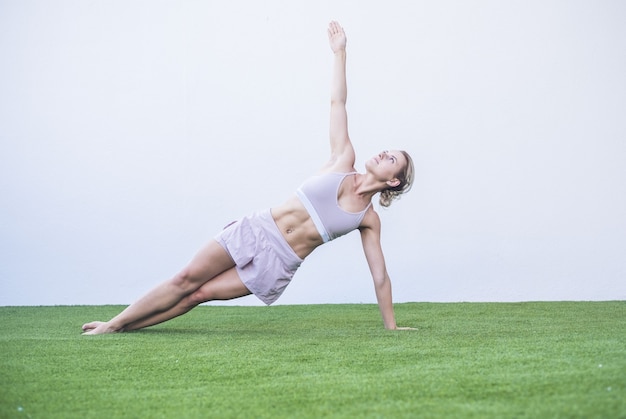 Beautiful young woman doing stretching exercise on grass in garden. Woman in sportswear balancing while doing exercise. Sporty young woman practicing yoga outdoors on green grass